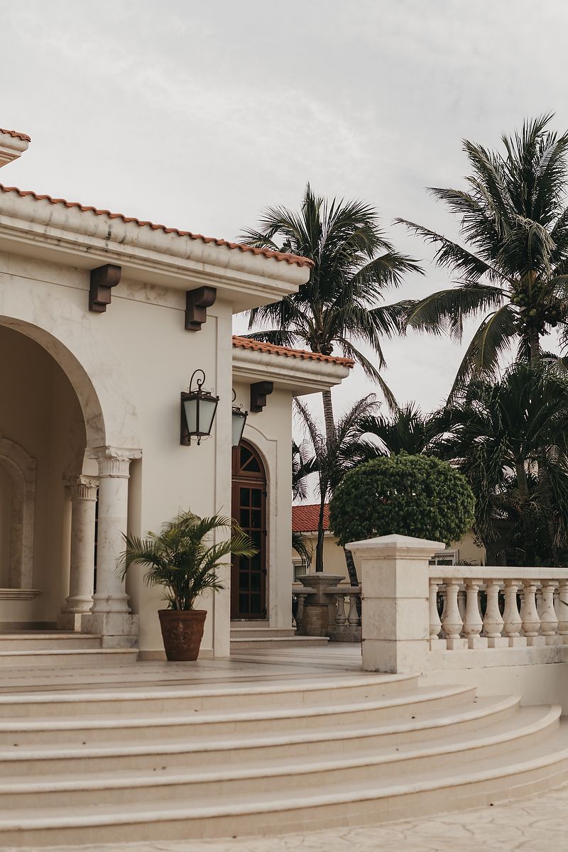 Mediterranean style white stucco exterior with curved archway, steps, balustrades and palm trees against overcast sky.