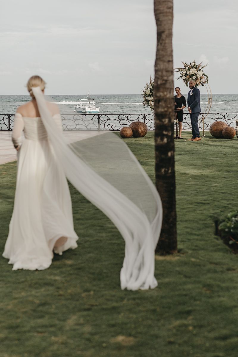 A flowing white wedding veil catches the ocean breeze on a grassy lawn with palm trees and sea views.