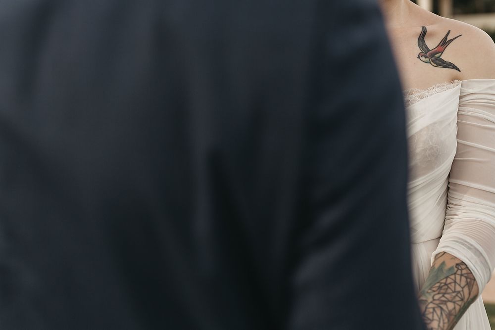Close-up detail of a bride's tattooed shoulder and white wedding dress against a dark suit.