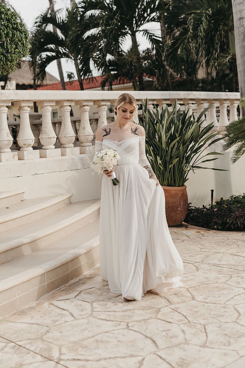 Elegant long-sleeve wedding dress with flowing skirt on white stone terrace surrounded by tropical palm trees.