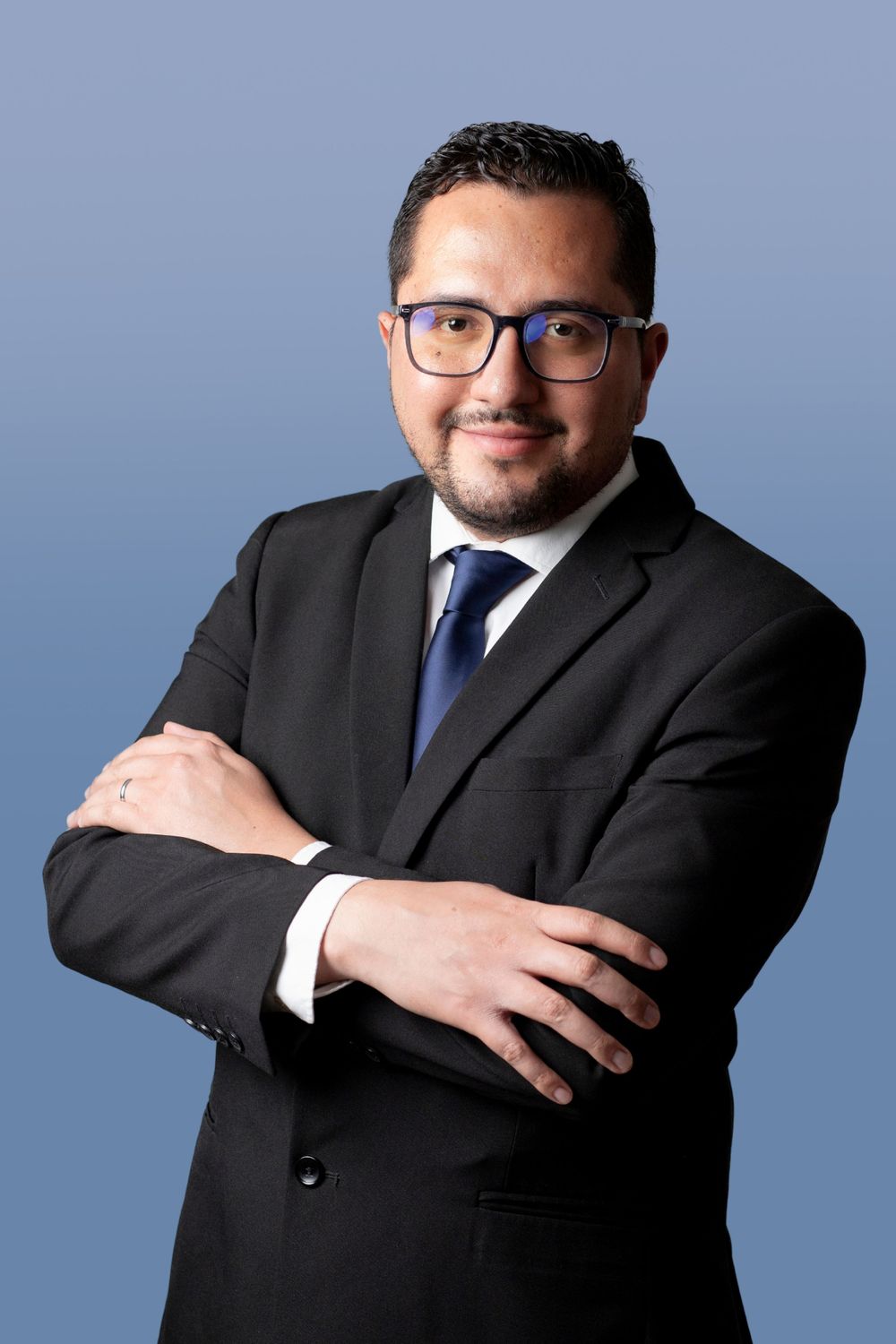 Professional business portrait of person in dark suit with glasses against blue background, arms crossed confidently.