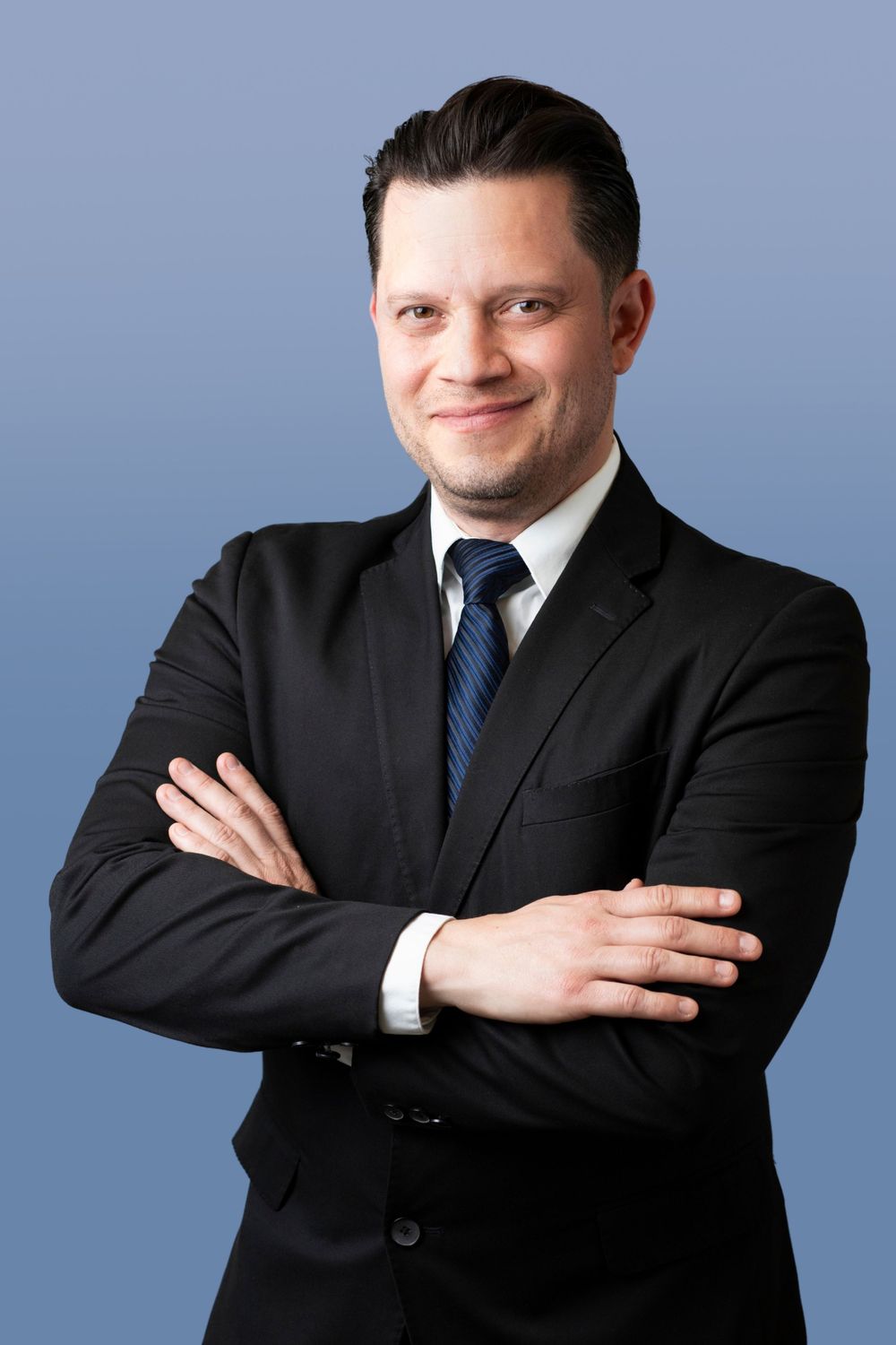 Professional wearing black suit and blue tie poses with crossed arms against blue background in studio portraits.