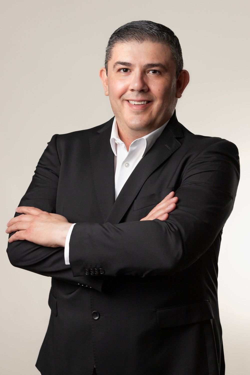 Professional business headshot of person in black suit jacket and white dress shirt with arms crossed against light background.