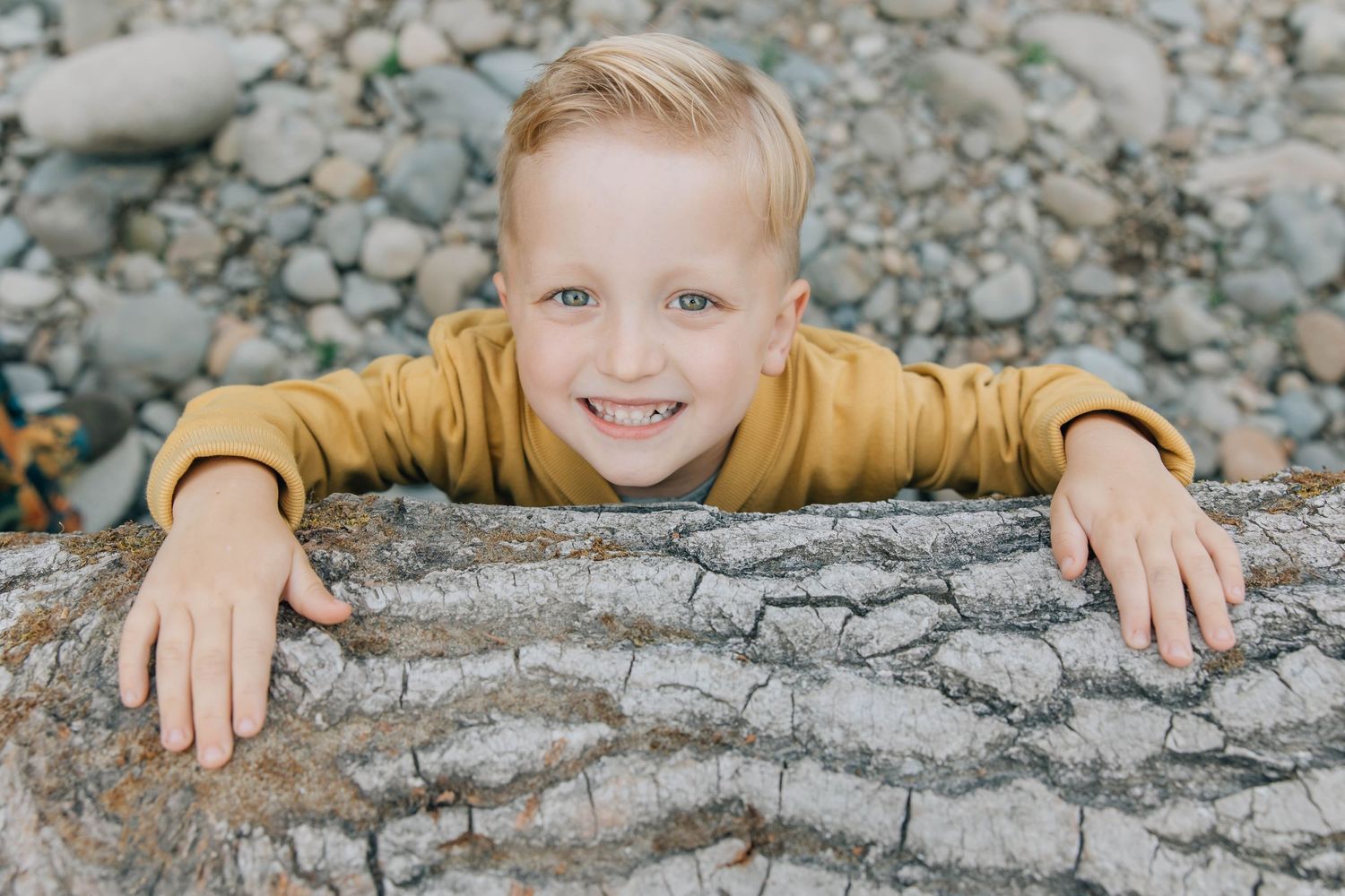 Happy young boy with blonde hair smiling while leaning on a large rock outdoors.
