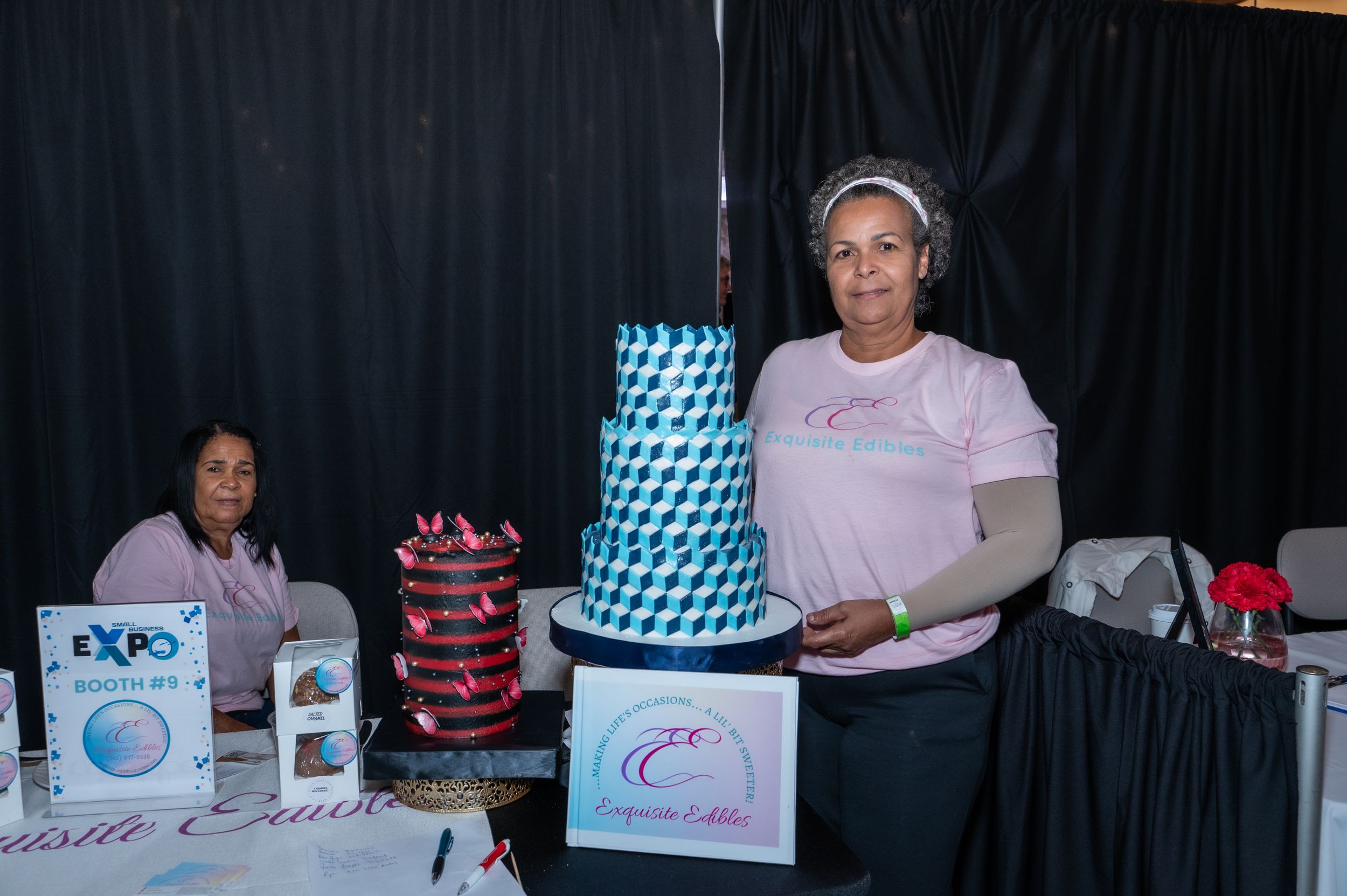 Woman proudly displays a blue geometric tiered cake at an expo event booth with pink branding signage.