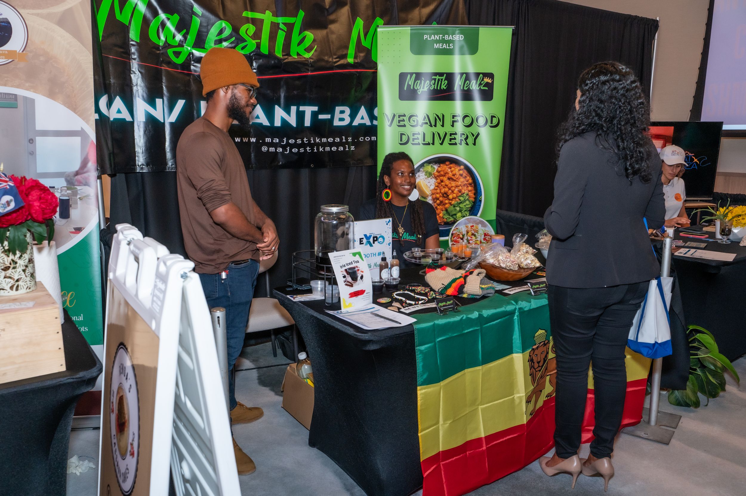 Vegan plant-based food vendor booth at an expo with colorful Rastafarian flag tablecloth and attendees browsing.
