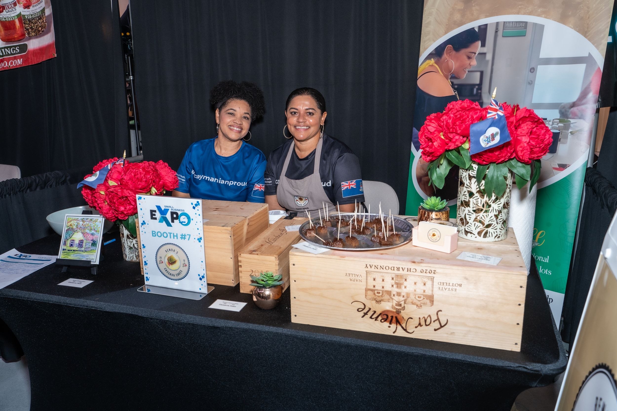 Two smiling women pose at a decorated expo booth with red flowers and a wooden display box.