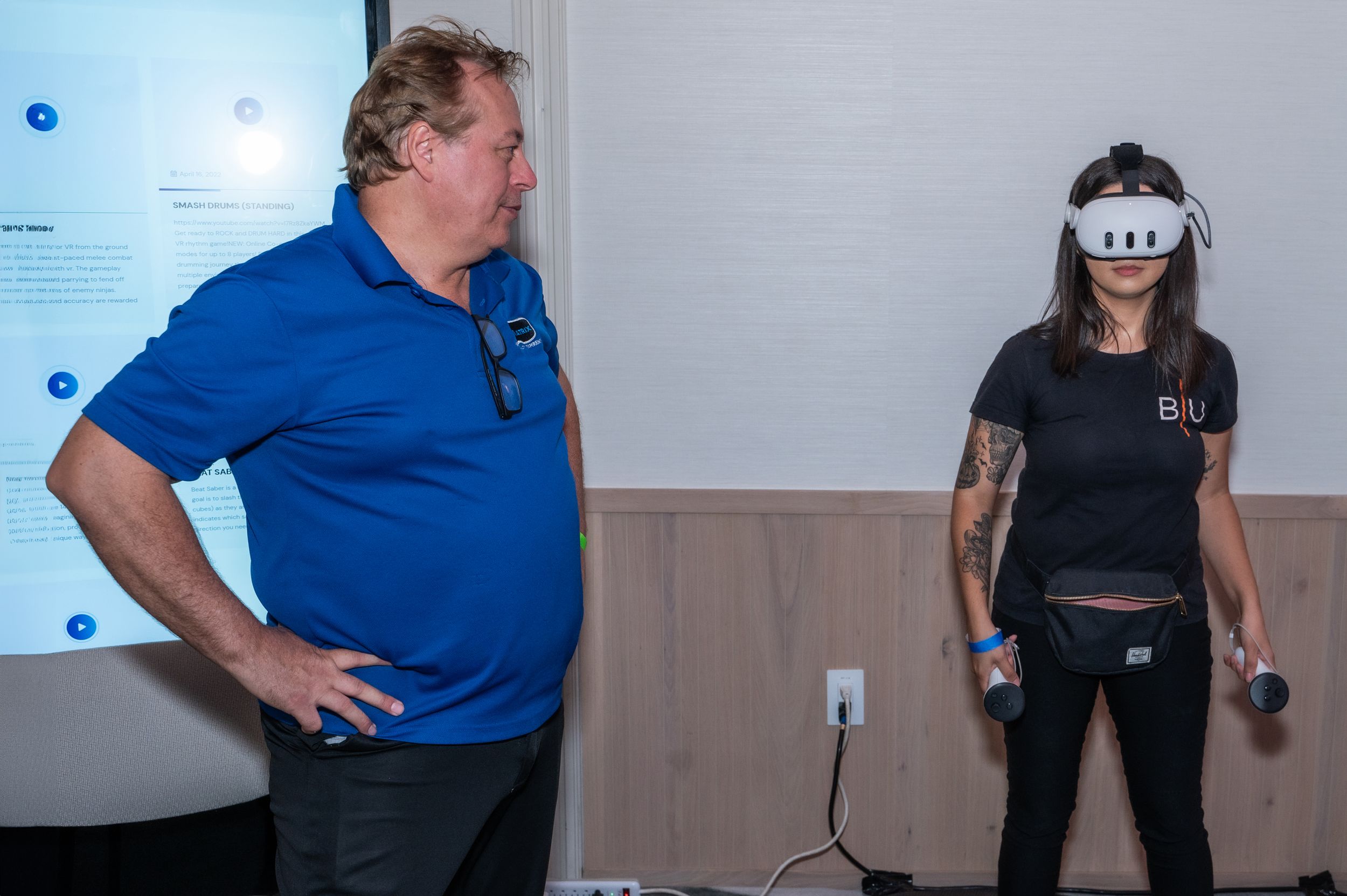 Man in blue polo watches woman using VR headset and controllers at a technology demonstration event.