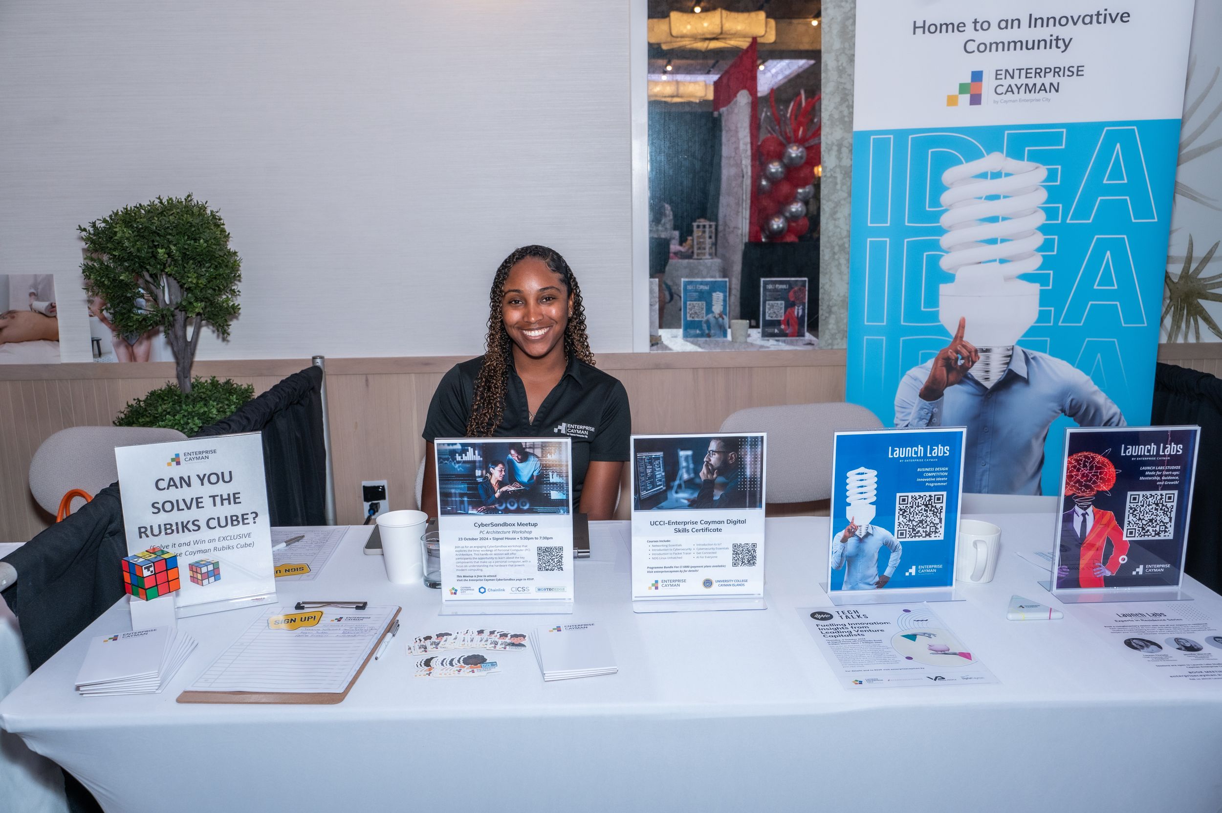 Young woman smiling at an innovation expo booth displaying IDEA signage and interactive displays at a business event.