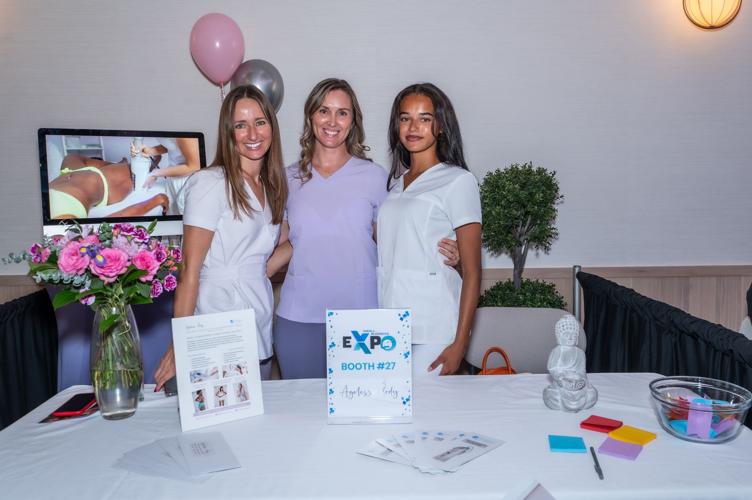 Three women smile at an Expo booth display with flowers, balloons, and promotional materials at an indoor event.