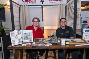 Two employees smile at a Custom Interiors and Tomlinson Furniture trade show booth display table.