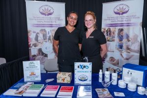 Two women smile at an expo booth displaying skincare products and promotional materials at a trade show.
