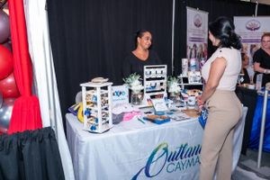 Two women at a Cayman Islands trade show booth displaying local products on a decorated table.