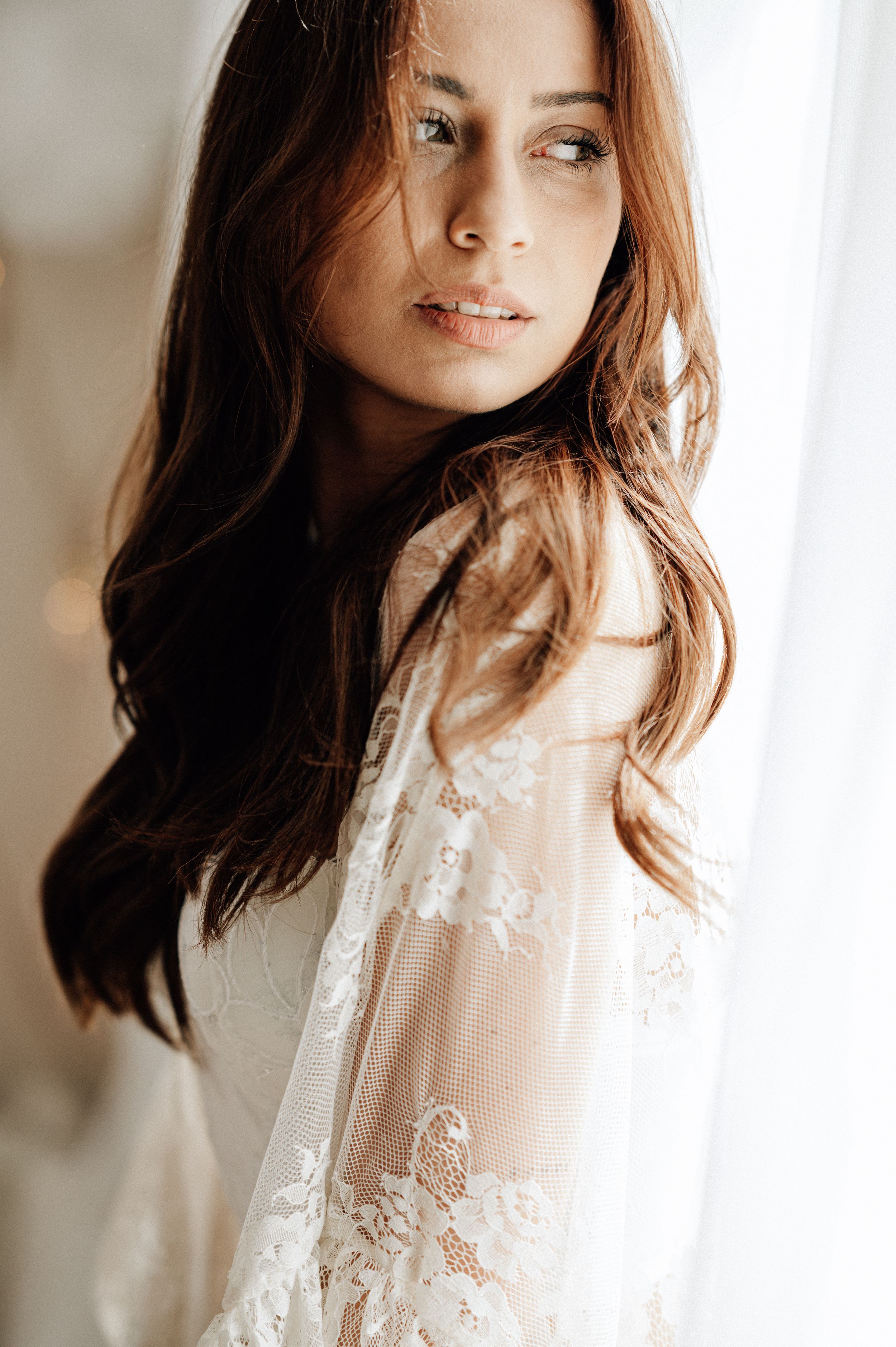 A person with flowing auburn hair wears a delicate white lace robe in a soft, ethereal boudoir setting.