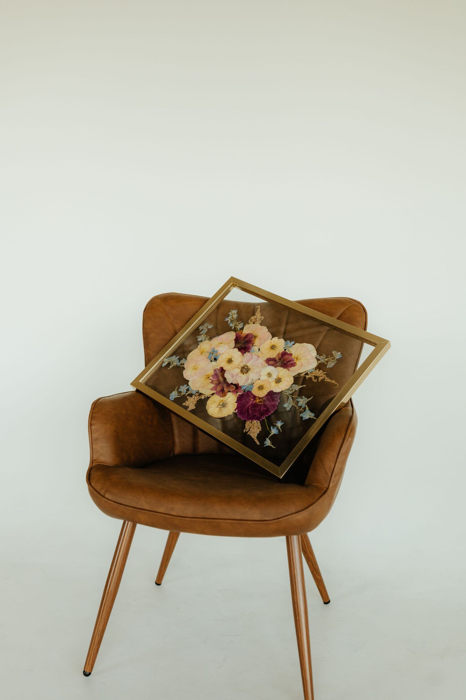 Pressed flower art in gold frame displayed on a brown velvet chair against white background.