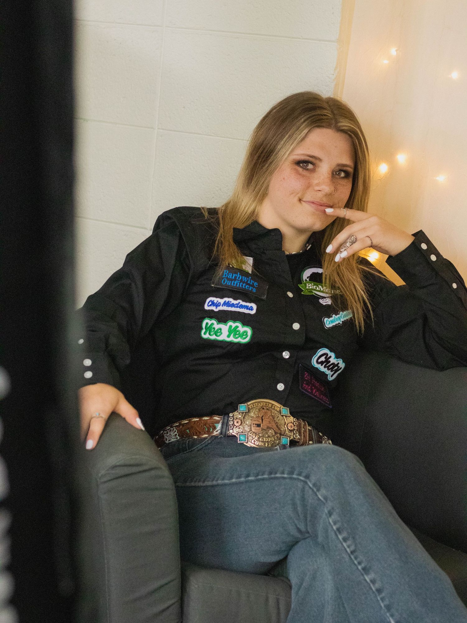 A person wearing a black jacket with sponsor patches and decorative belt sits comfortably on a couch with string lights in background.