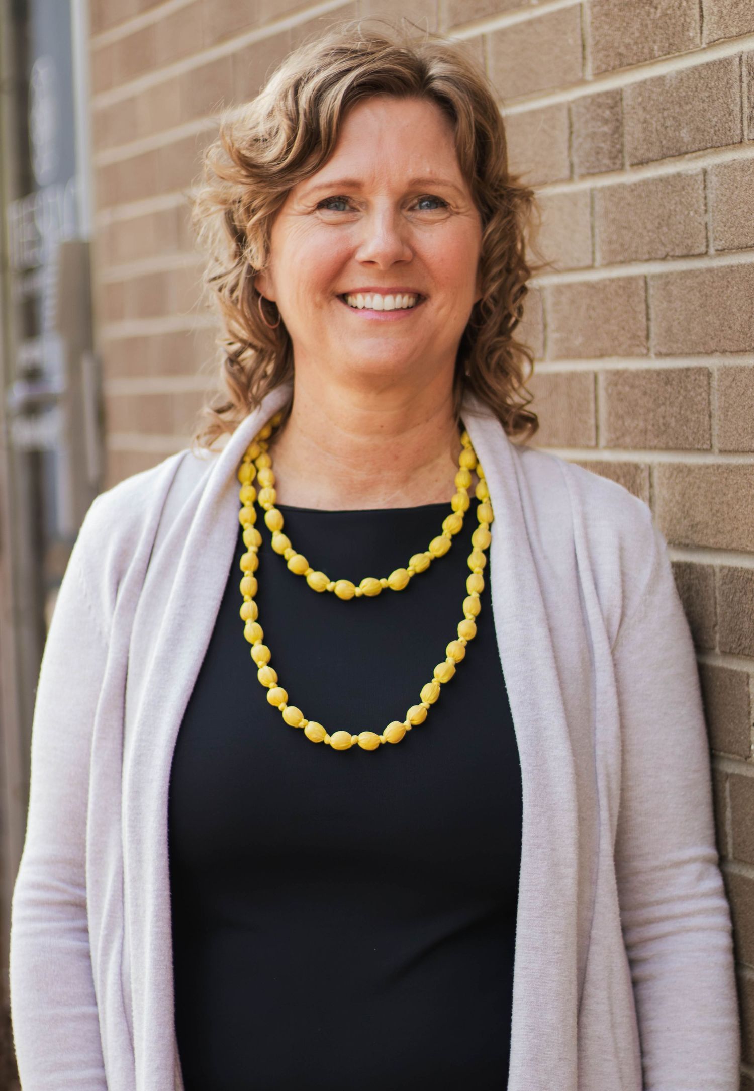 Professional portrait featuring beige cardigan and vibrant yellow beaded necklace against brick wall.