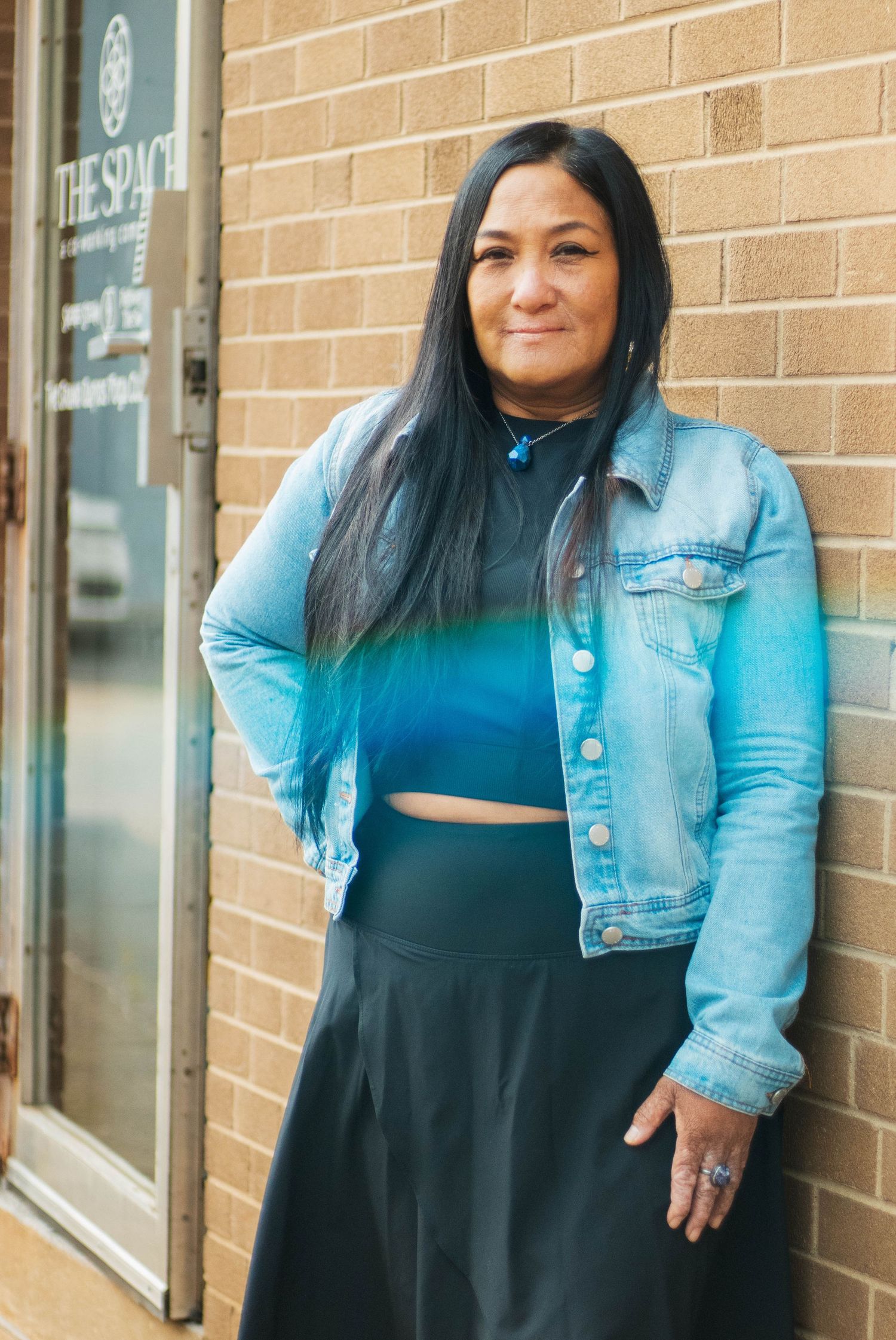Woman in denim jacket and black skirt poses confidently against brick wall outside store.