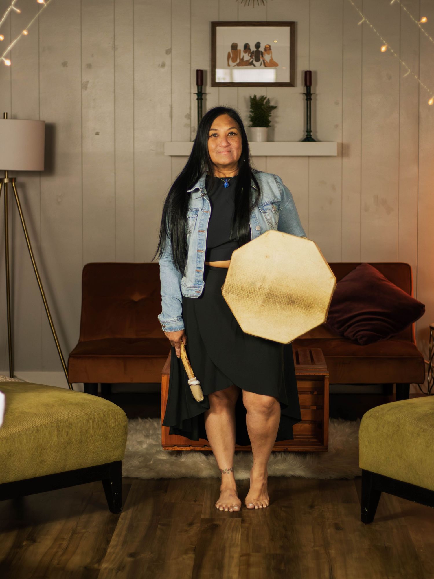 Long-haired woman in denim jacket holds decorative item in cozy room with string lights.