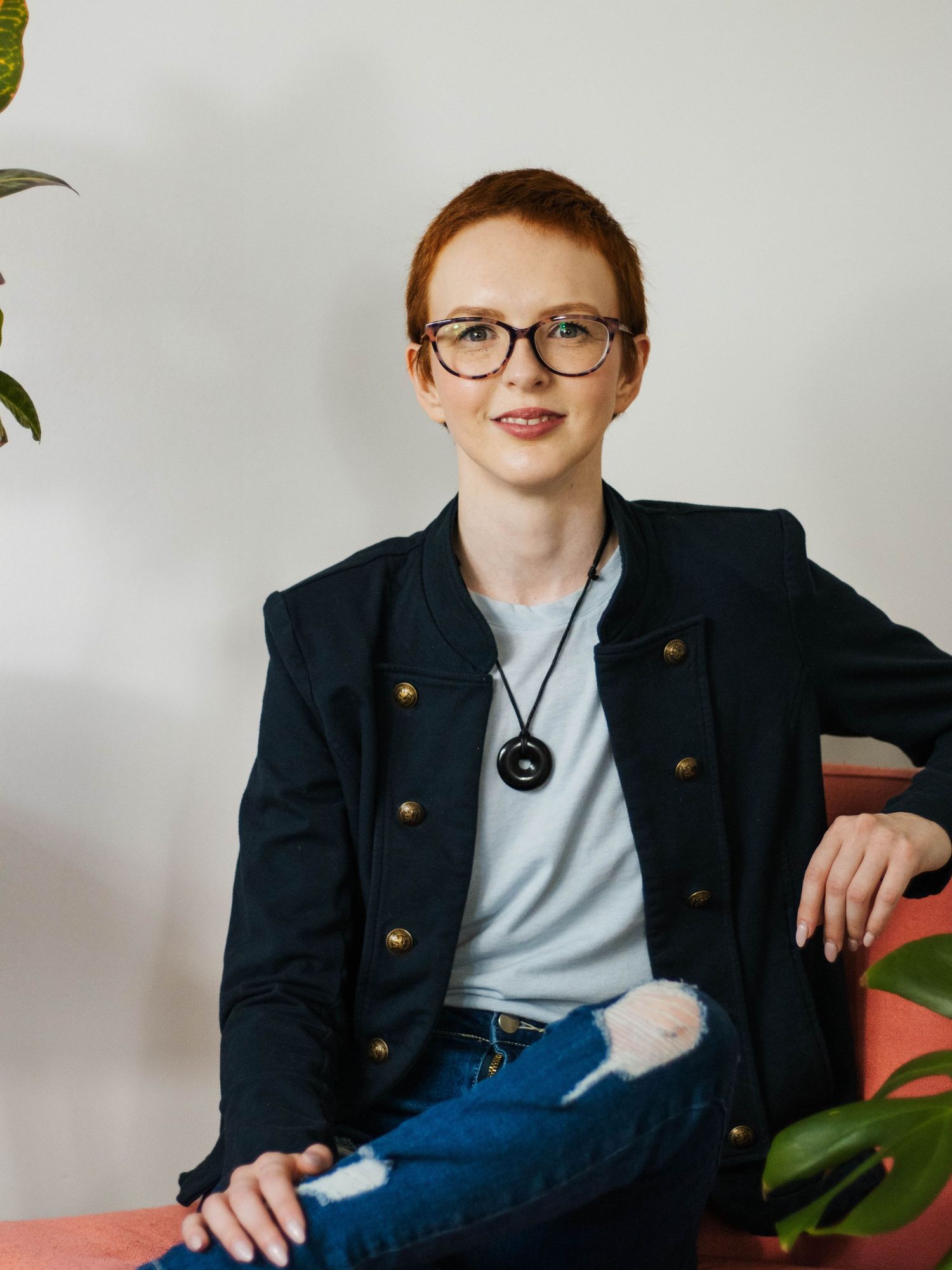 Professional wearing glasses and navy blazer with brass buttons sits casually in a modern studio setting.