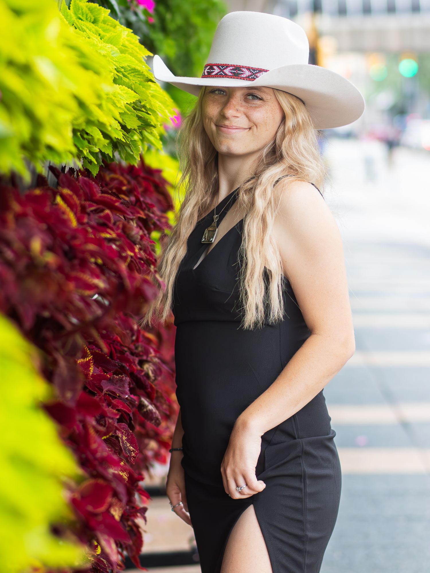 A person in a white cowboy hat and black dress poses next to bright red and green foliage on an urban street.