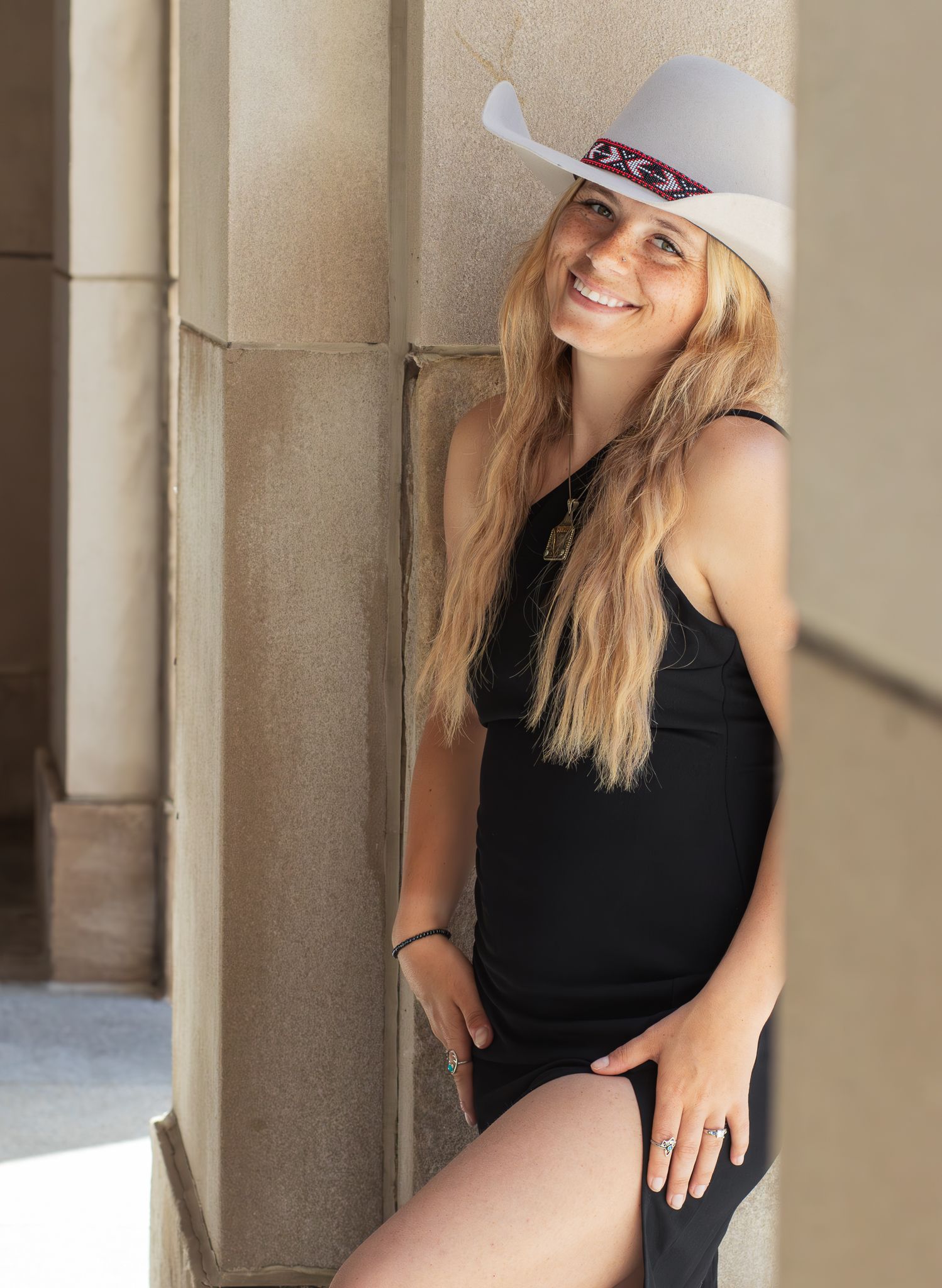 A person in a white cowboy hat and black dress leans against a stone column while smiling in natural lighting.