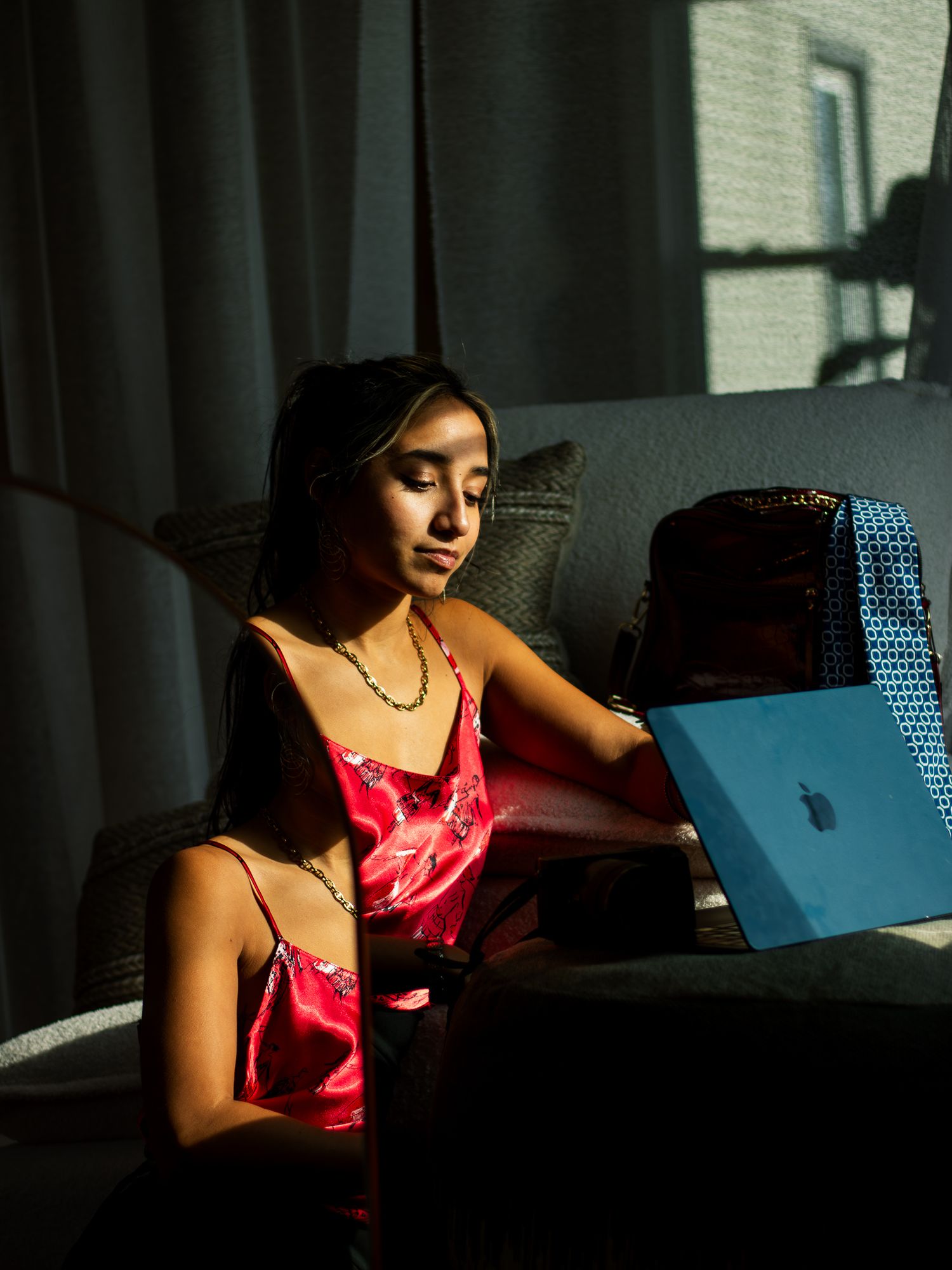 Someone in a red silk camisole works on a blue laptop while sitting in moody window lighting.