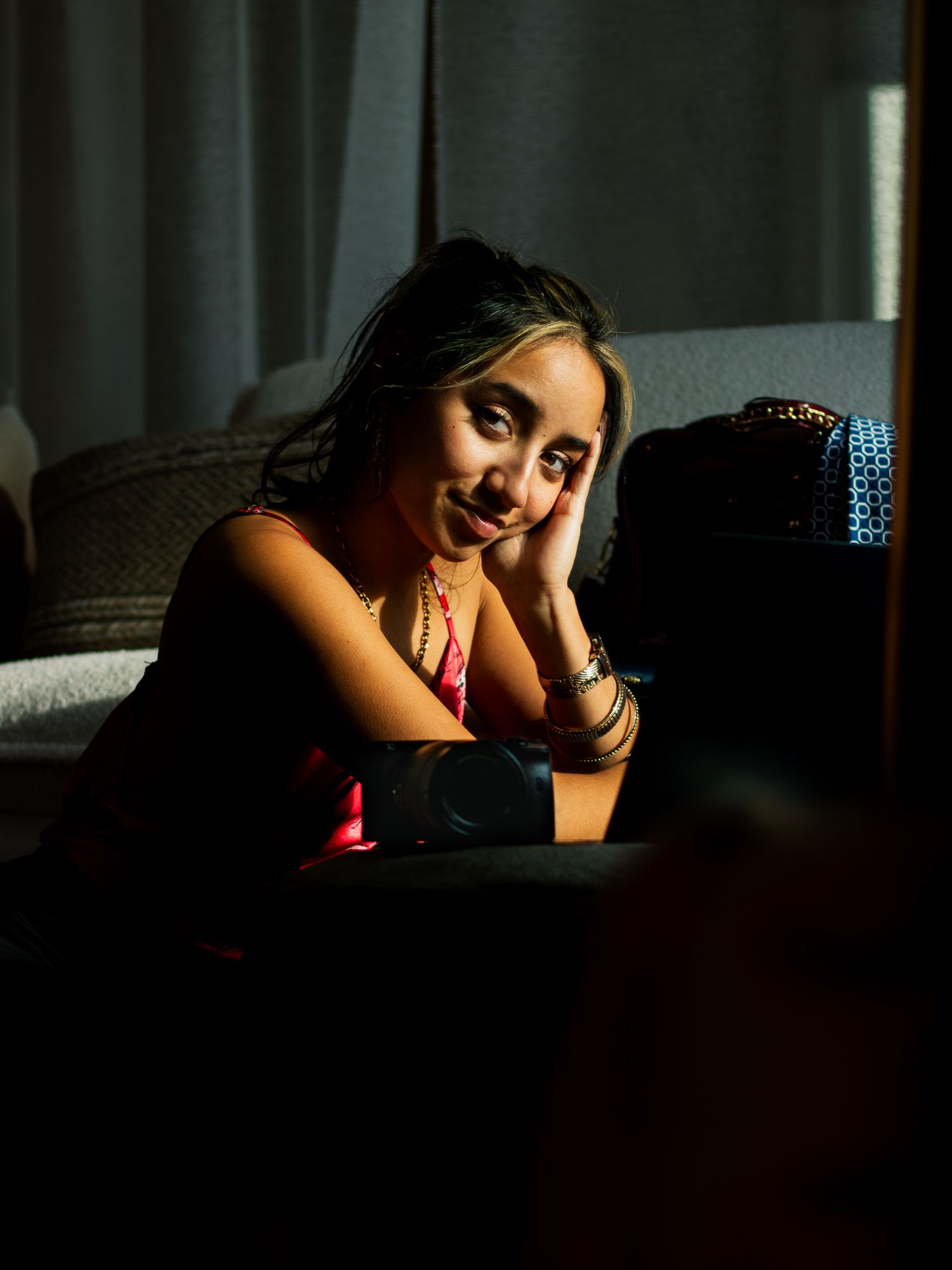 A dramatic portrait in moody lighting shows someone in a red top resting their head on their hand while seated on a couch.