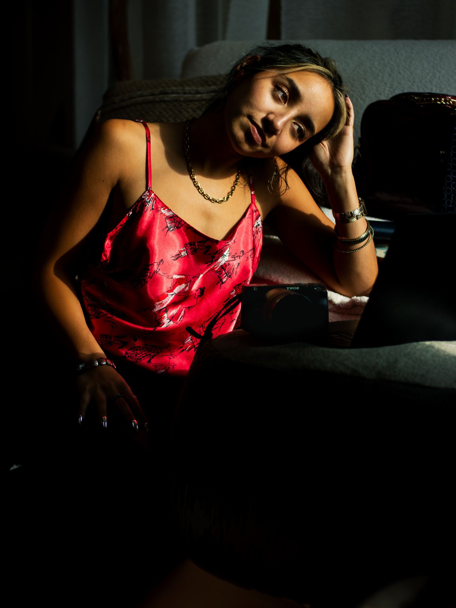 Dramatic moody portrait shows a person in a red silk camisole lounging against dark furniture in low light conditions.