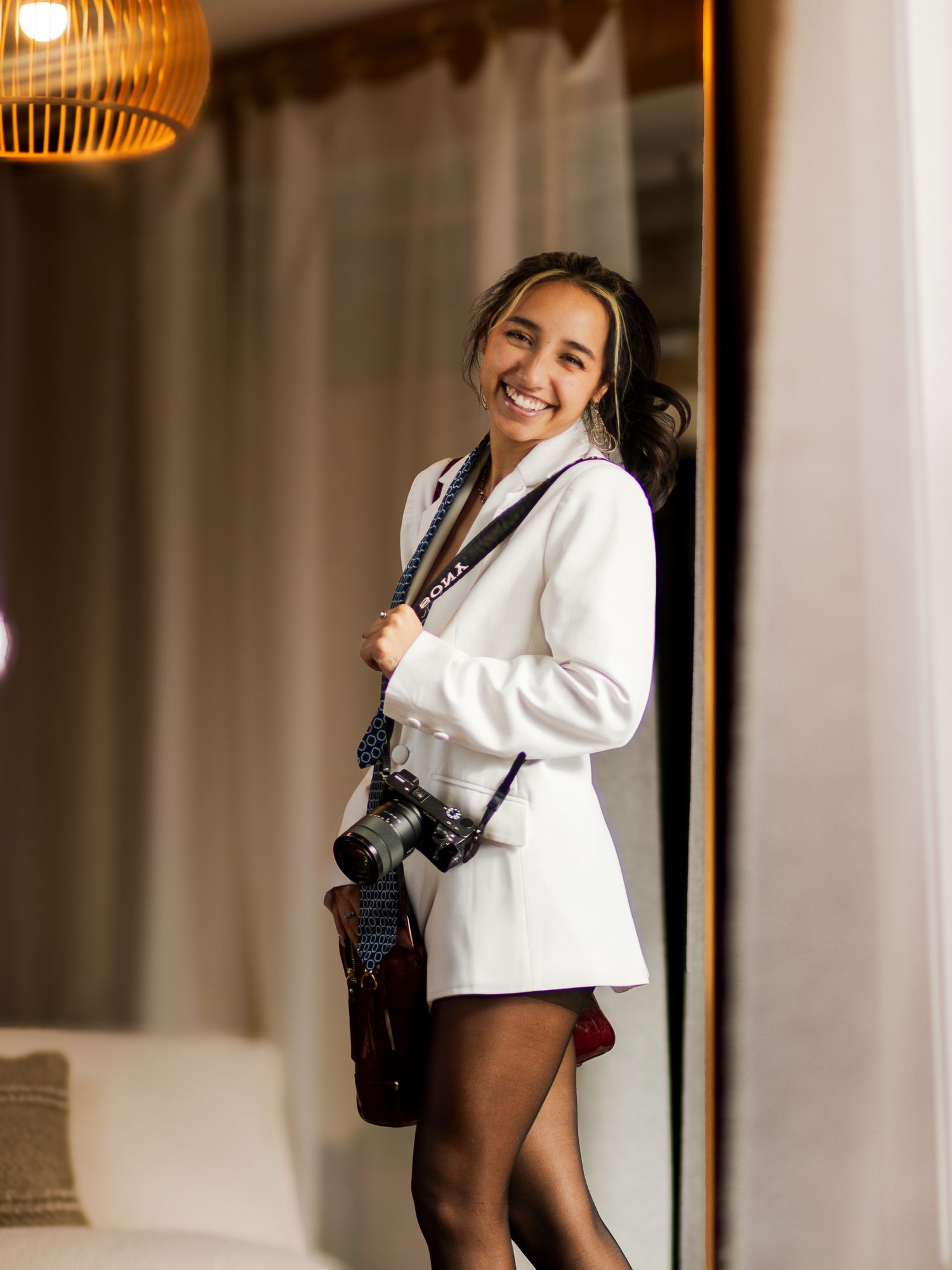 Professional photographer in white blazer and black shorts poses with camera in warm interior setting.