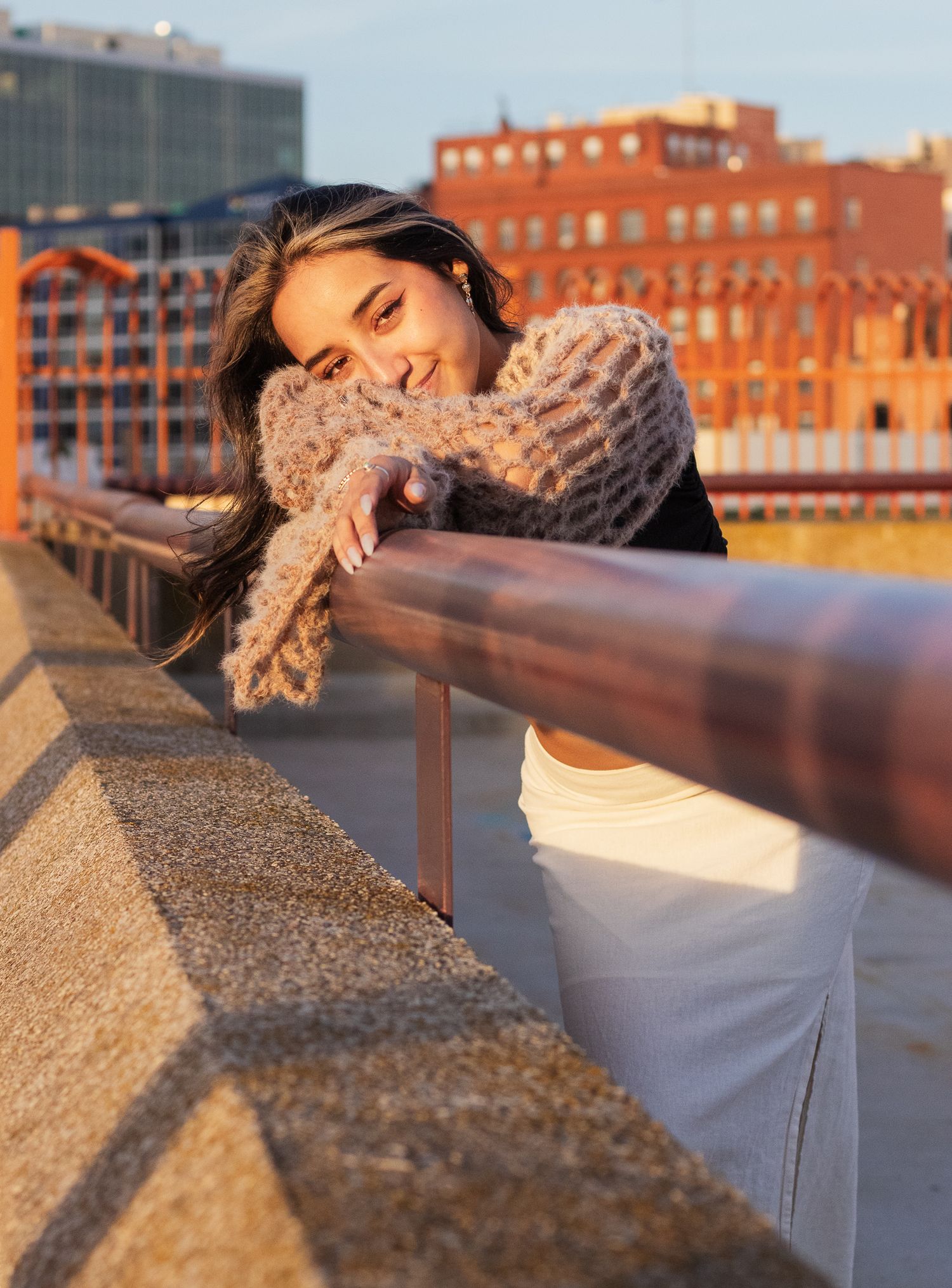 Urban fashion portrait with model leaning on railing at sunset with city buildings in background.