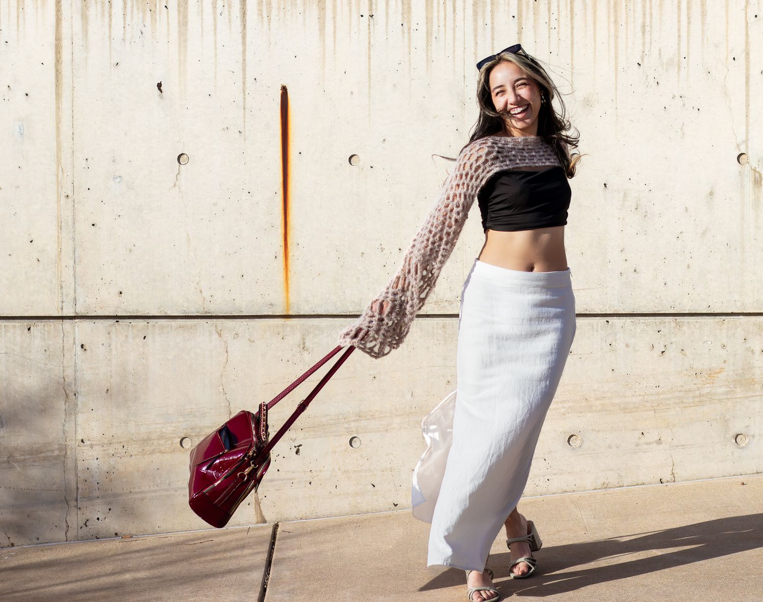 Fashion model swinging burgundy bag while posing against concrete wall in stylish outfit.