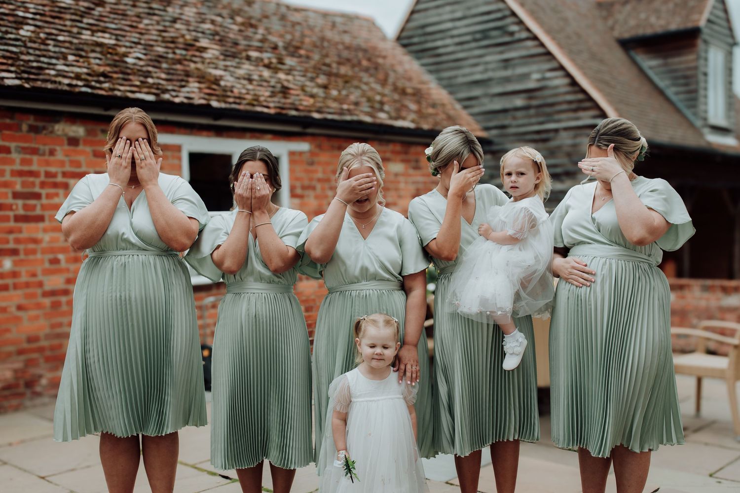 Group in matching sage green dresses pose playfully at a rustic brick venue. Houchins essex