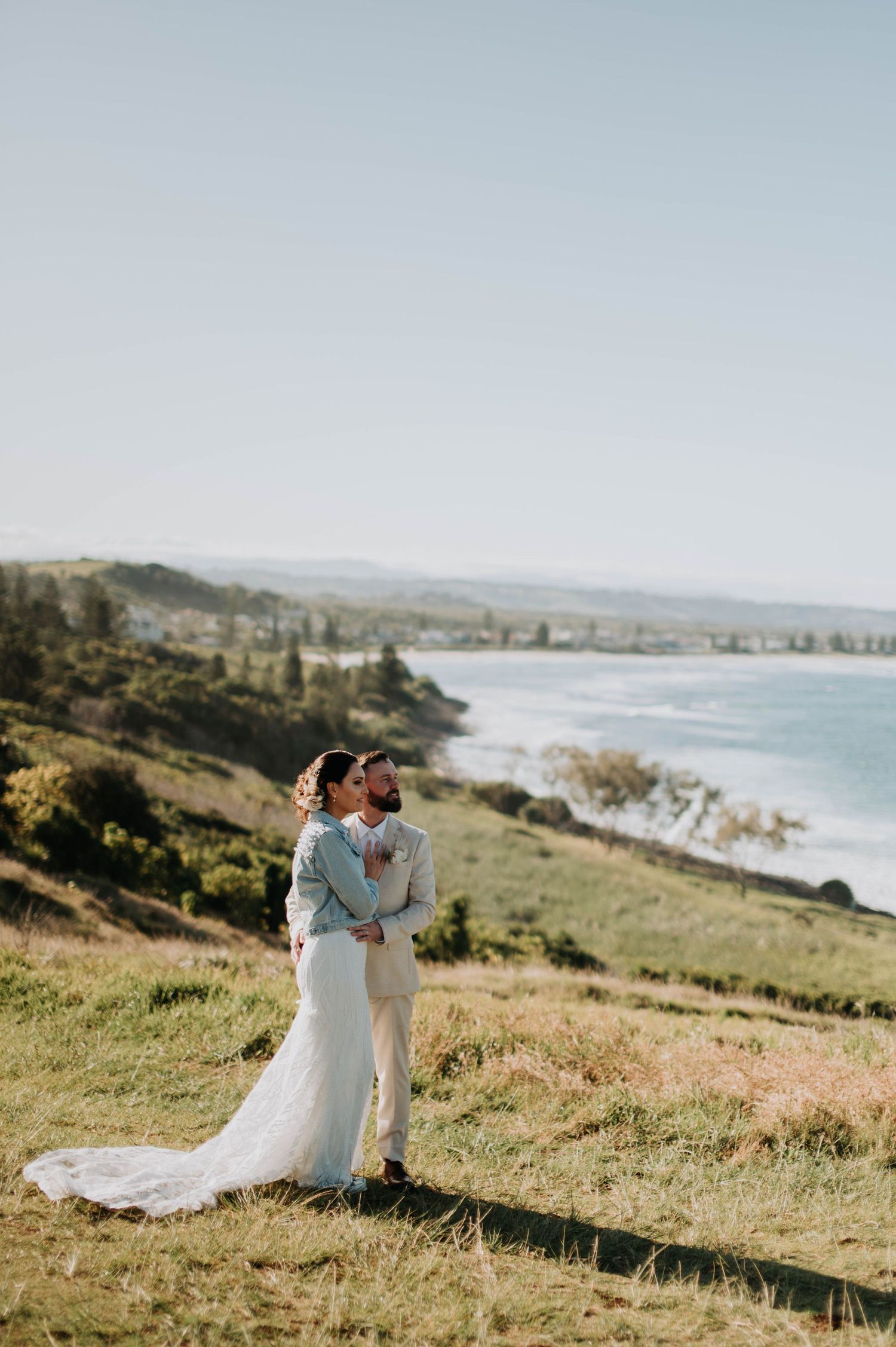 Newlyweds embrace on a coastal hillside with stunning ocean views and rolling landscape.