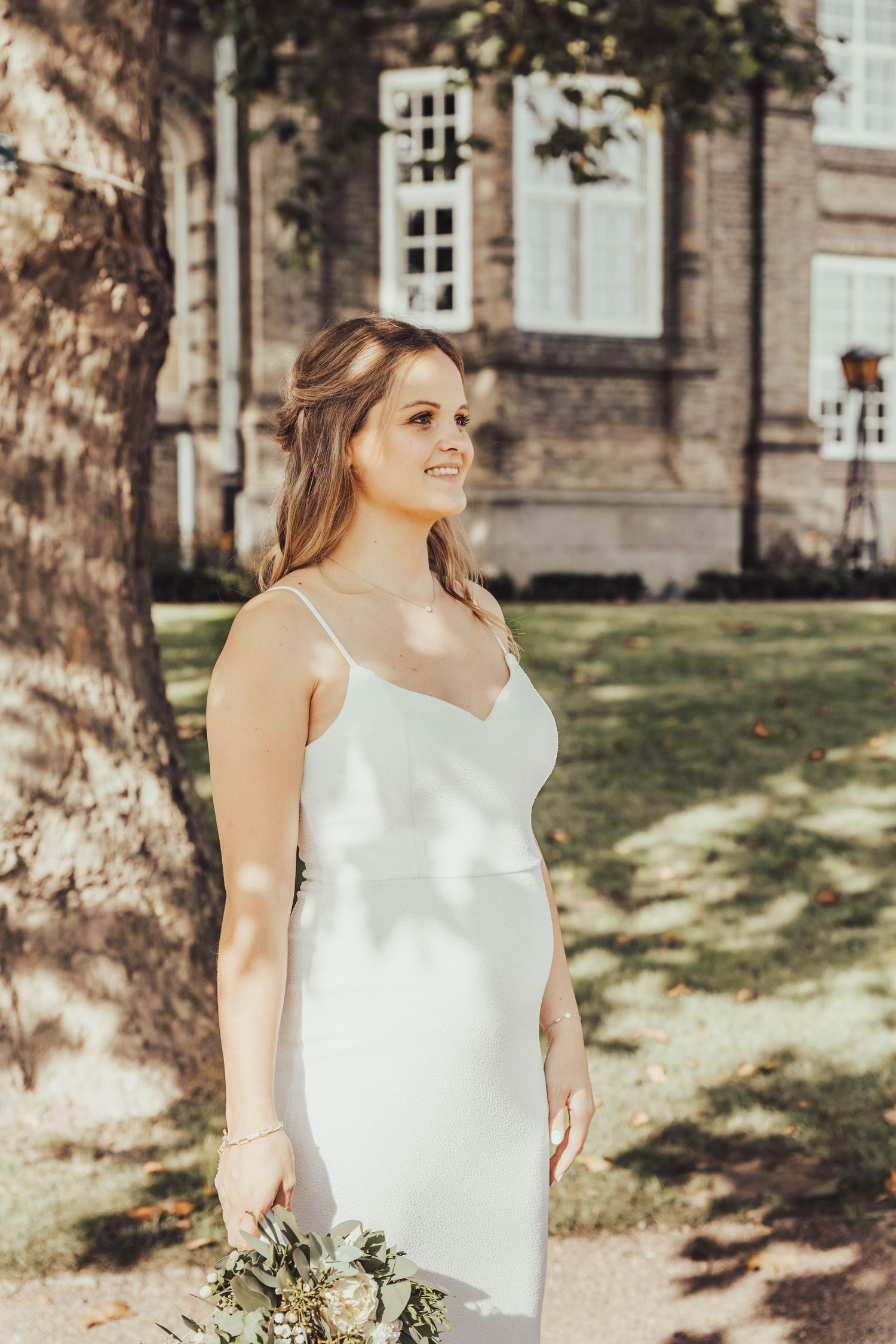 A bride in a sleek white gown poses outdoors beside a historic stone building with dappled sunlight.