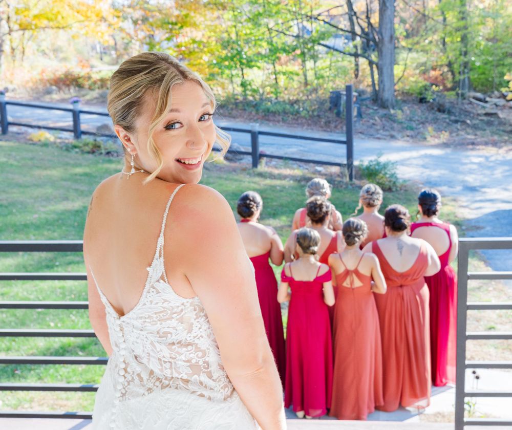 Bride in lace dress looks back at bridesmaids lined up in red and coral dresses outdoors.
