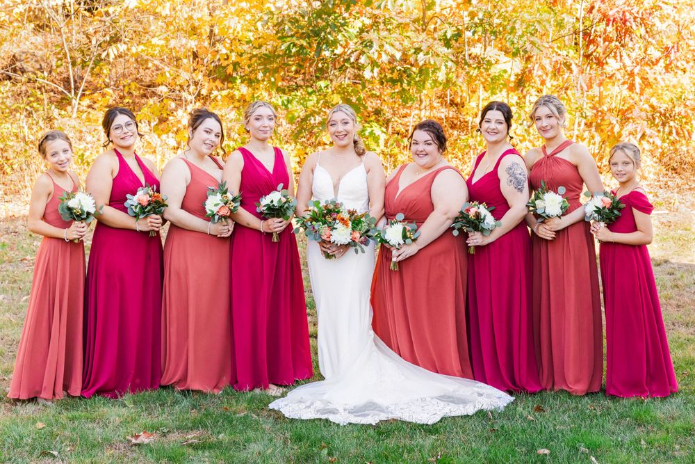 Wedding party poses outdoors in shades of red and burgundy dresses against fall foliage.