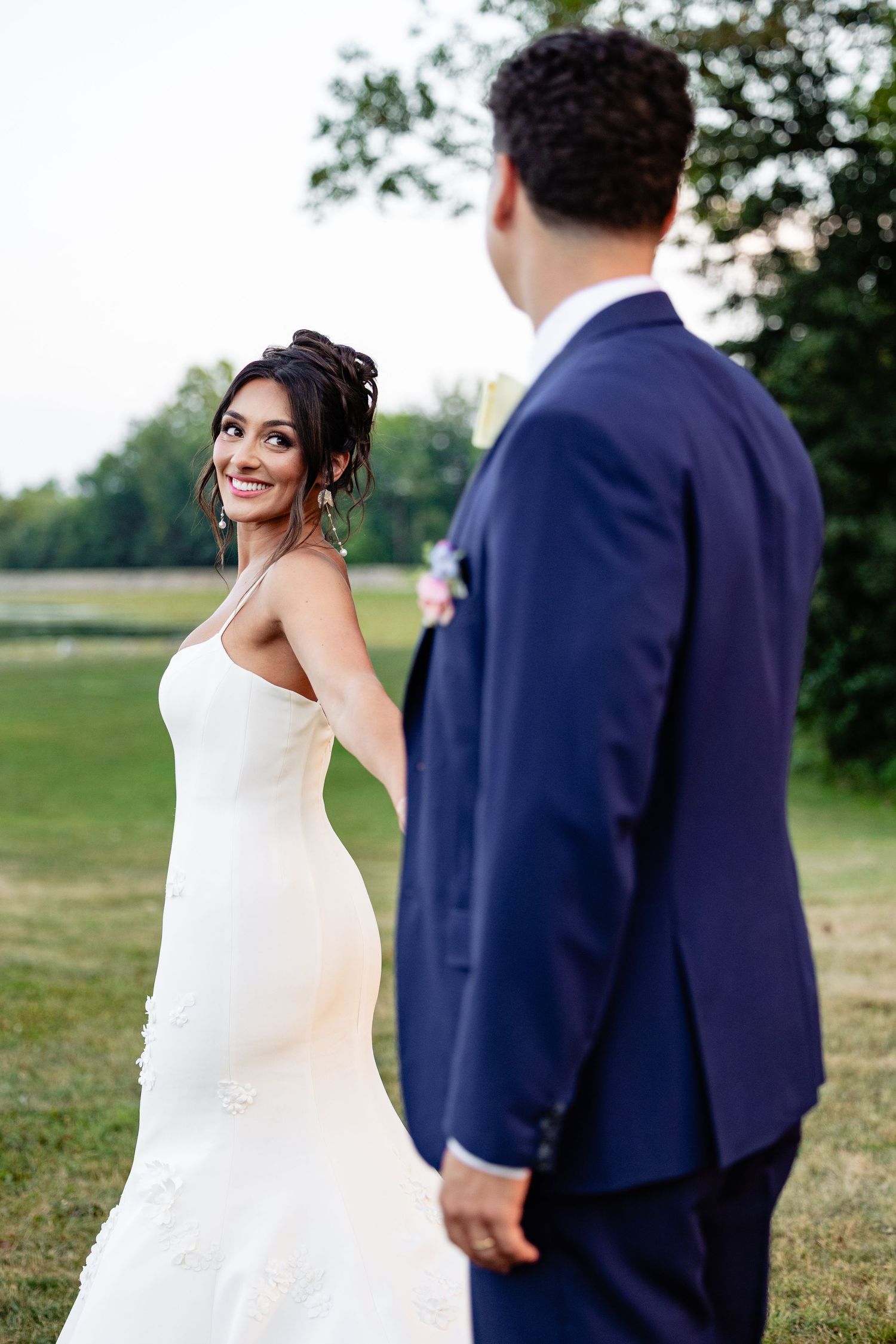 Smiling bride in a sleek white gown looks back while holding hands with her groom outdoors.