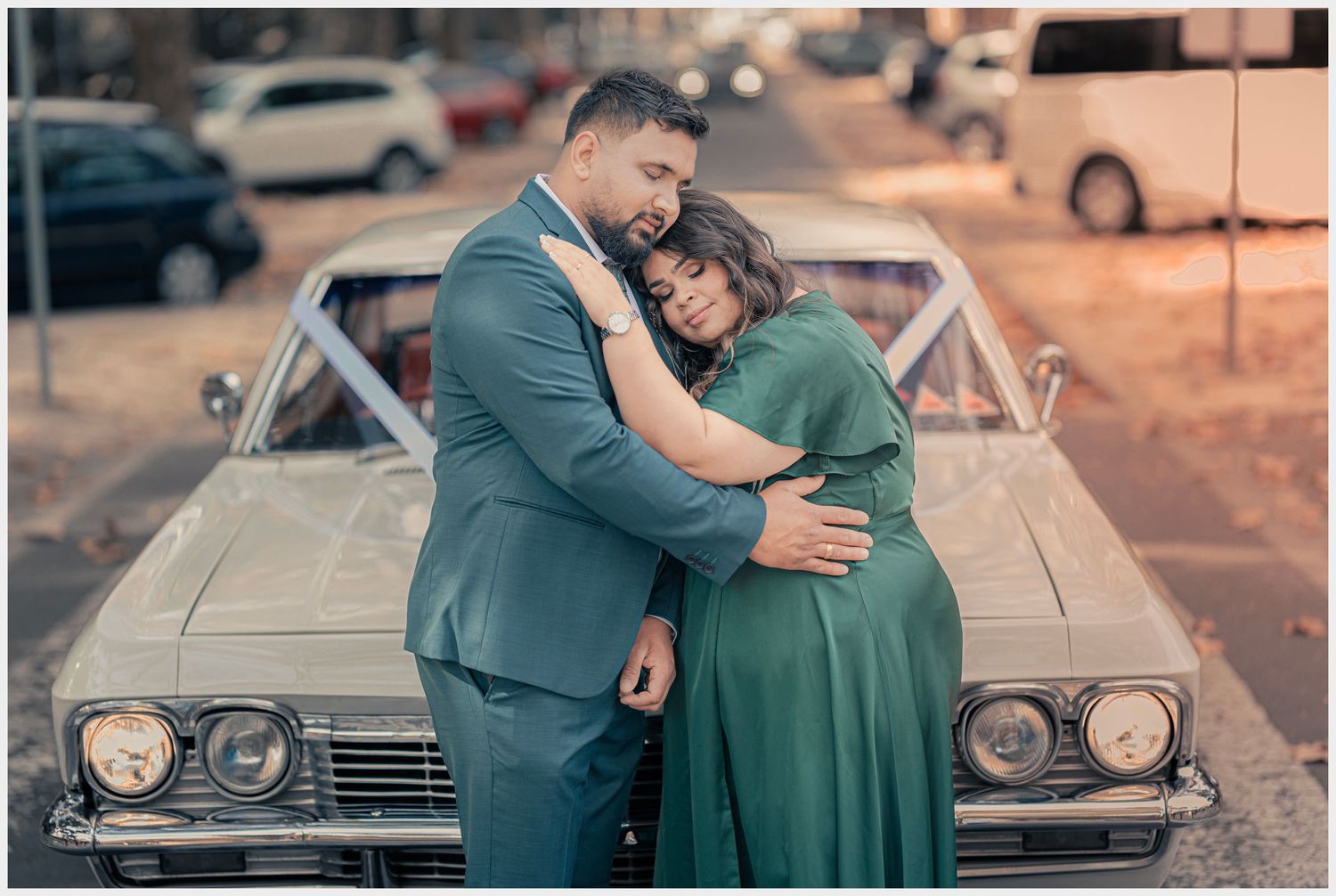 Couple embracing in formal wear against vintage car at sunset.