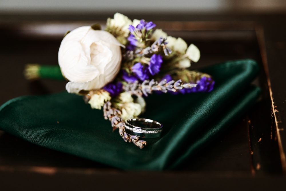 Wedding boutonniere with white rose and purple flowers resting on emerald green fabric.