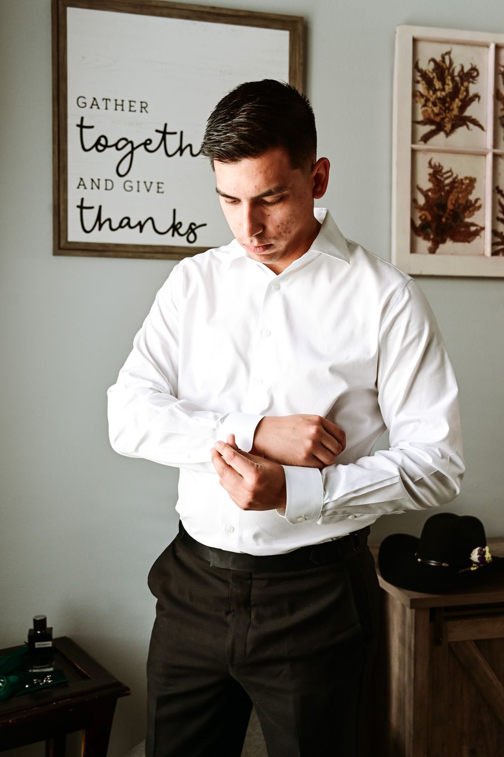 Person adjusting white dress shirt cuffs near wall art that reads 'Gather together and give thanks'.
