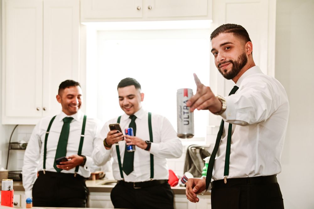 Groomsmen in white shirts and green suspenders sharing drinks during wedding preparations.