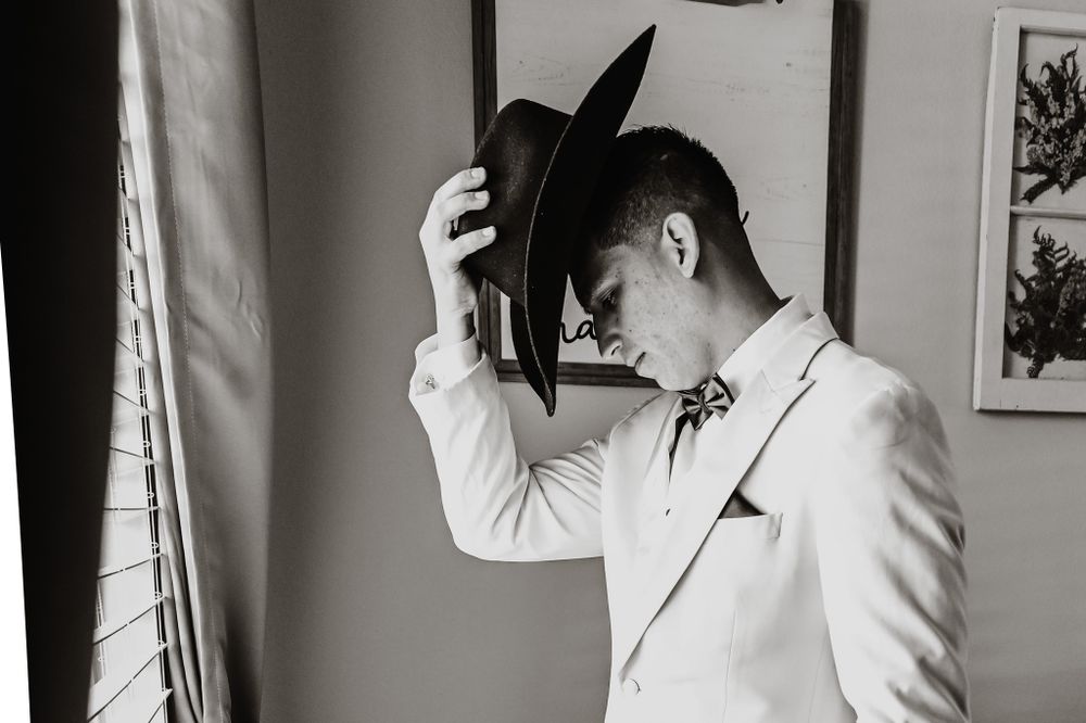 Groom in white suit jacket adjusts his hat while standing by window in black and white photo.
