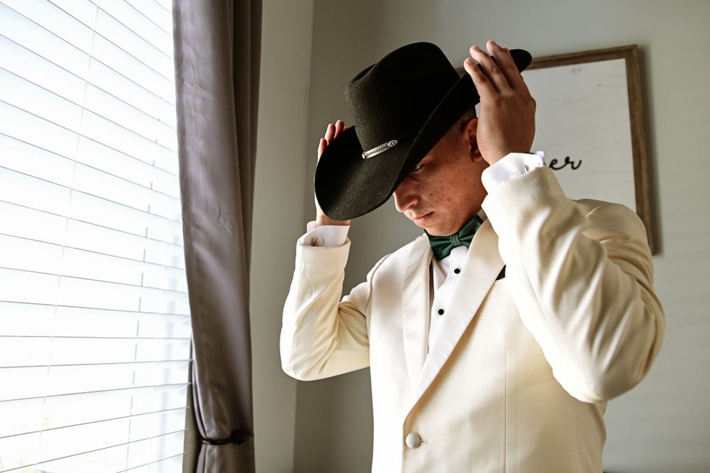 Side view of groom in white tuxedo adjusting black hat by window.