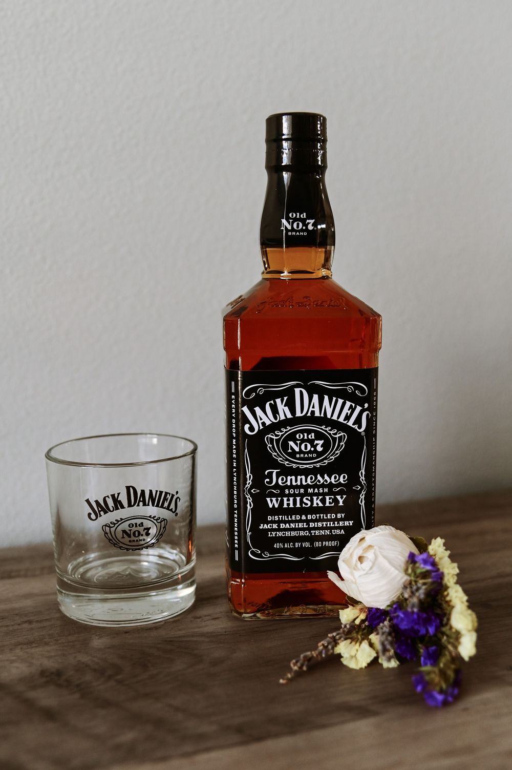 Jack Daniels whiskey bottle and glass with purple flower boutonniere on wooden surface.