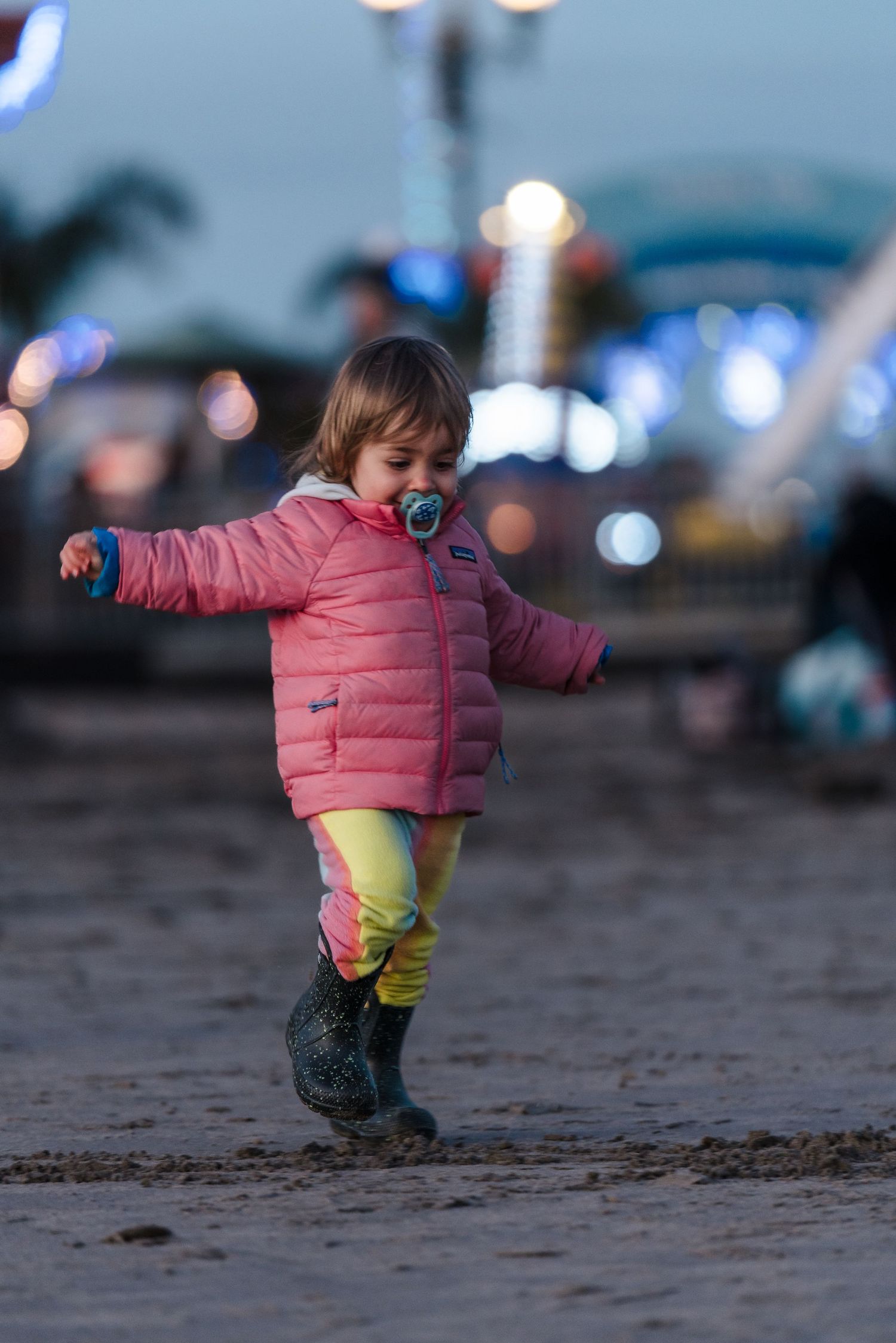 A small child in a pink puffy jacket plays on a beach at dusk.  Located near the Santa Cruz Wharf, California.