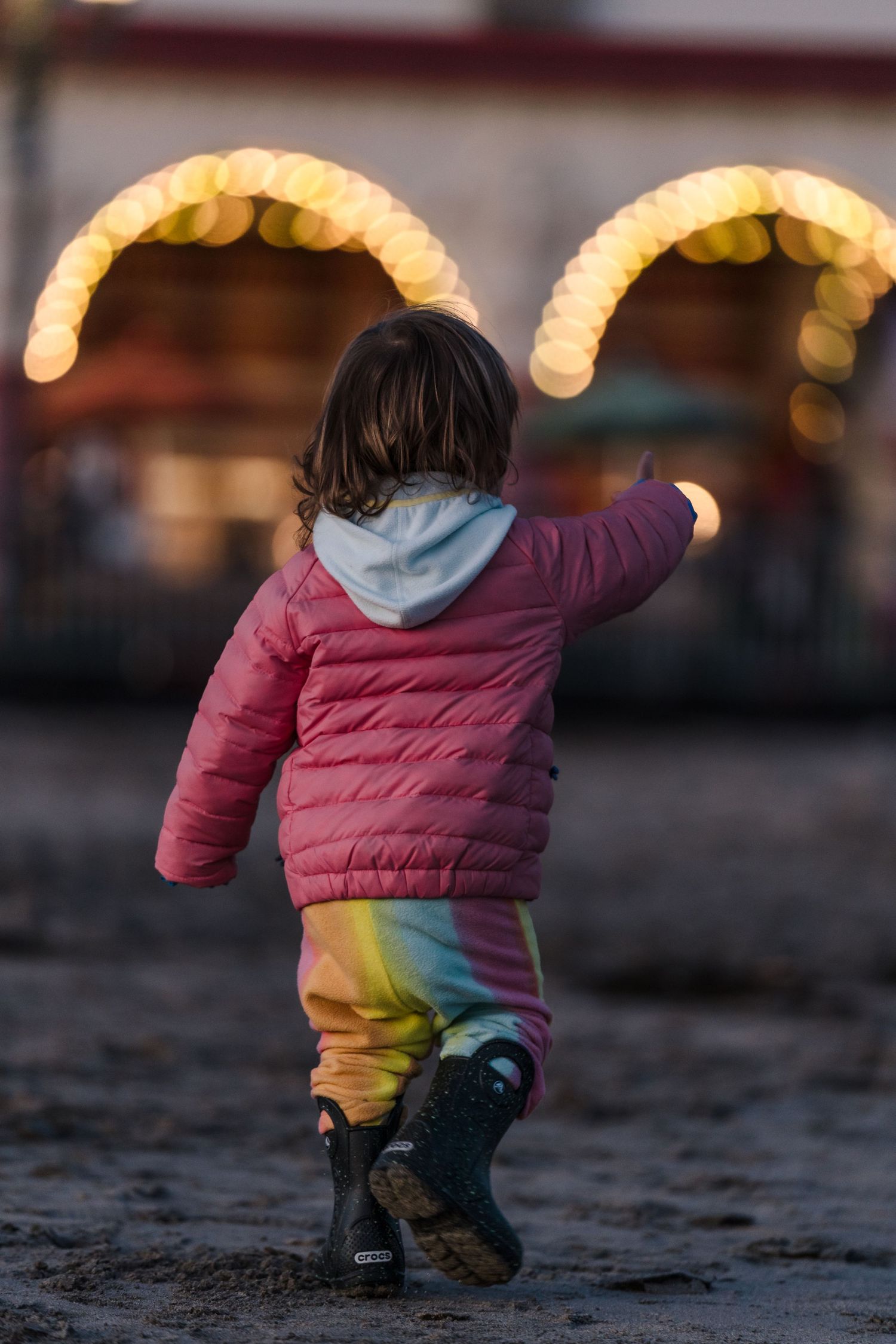 A child in a pink coat and rainbow pants walks away from glowing carnival lights.  Located near the Santa Cruz Wharf, California.