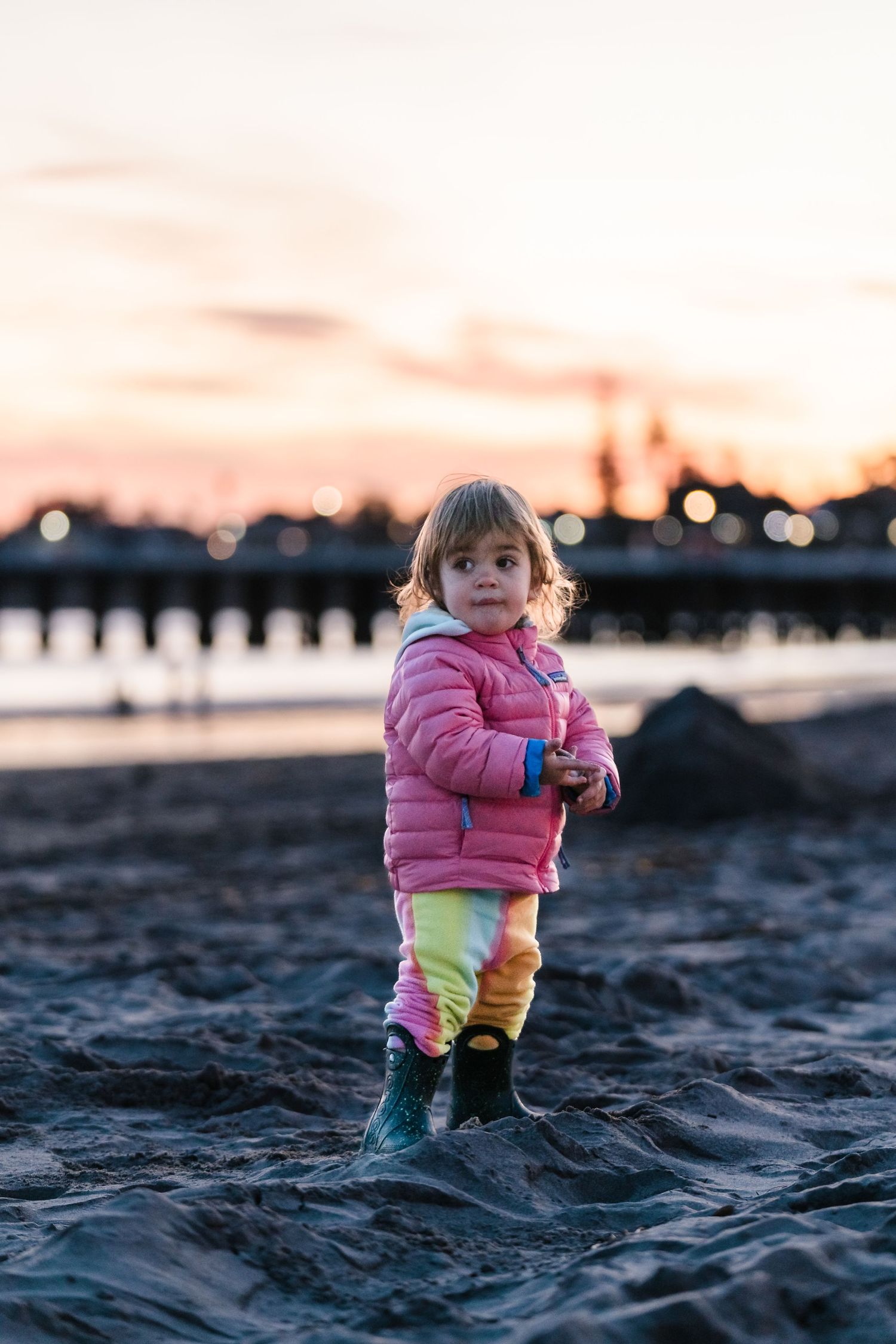 A small child in pink coat and yellow pants stands on a beach at sunset.  Located near the Santa Cruz Wharf, California.