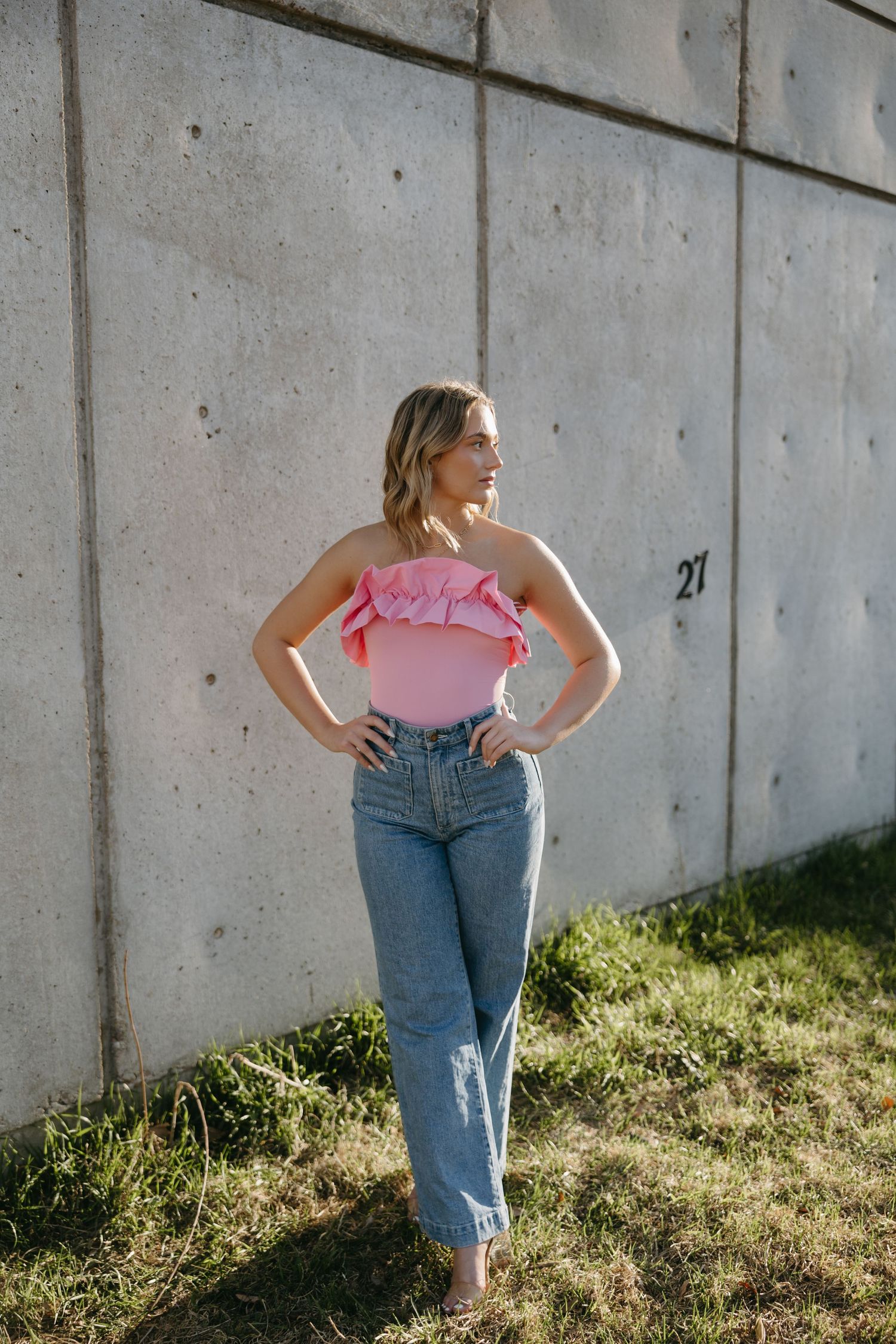Casual summer fashion featuring pink ruffled top and high-waisted distressed jeans against concrete wall backdrop.