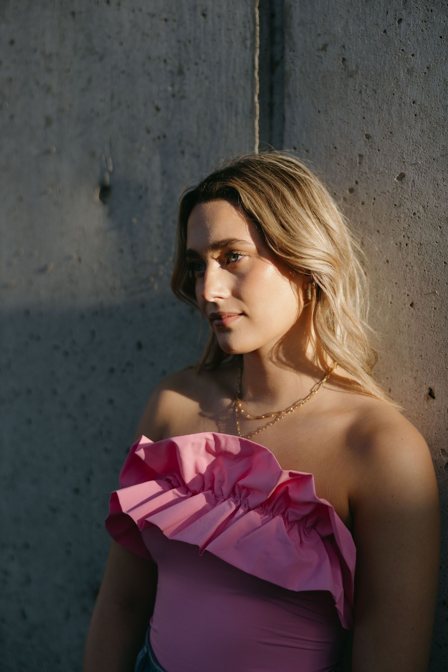 Portrait in dramatic lighting featuring pink one-shoulder ruffle top against concrete background.